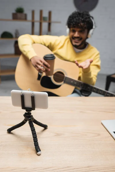 Smartphone en trípode cerca de podcaster indio borroso en auriculares que sostienen el café para ir y la guitarra acústica durante la corriente en el estudio de podcast, smartphone en trípode - foto de stock