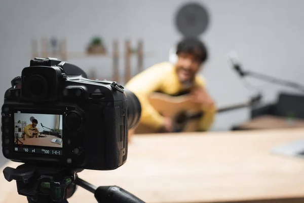 Screen of digital camera on tripod standing near blurred young indian podcaster holding acoustic guitar near microphone and laptop on table in podcast studio — Stock Photo