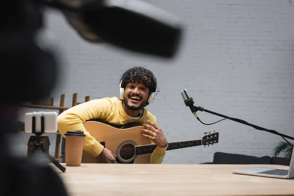 Позитивный индийский подкастер в наушниках, держащий акустическую гитару рядом с микрофоном, размытый смартфон на штативе и кофе, чтобы пойти на деревянный стол в студии — стоковое фото