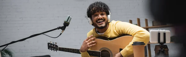 Cheerful indian podcaster in headphones holding acoustic guitar near smartphone on tripod, coffee and microphone during stream in studio, banner — Stock Photo
