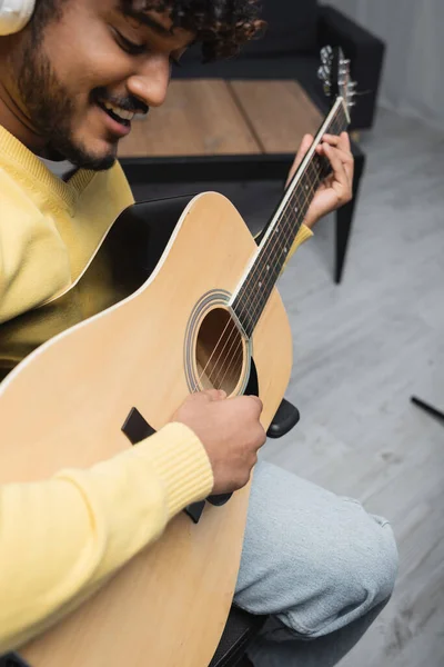 Размытый и улыбающийся молодой индийский музыкант в повседневной одежде и беспроводных наушниках, поющий и играющий на акустической гитаре во время выступления в студии — стоковое фото