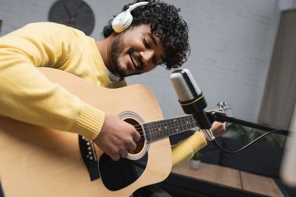 Positivo y joven podcaster indio en auriculares tocando la guitarra acústica cerca del micrófono en el stand durante la transmisión en estudio podcast - foto de stock