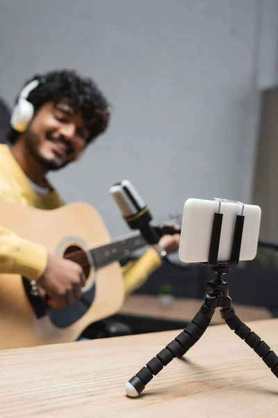 Smartphone en trípode en la mesa cerca de podcaster indio borroso en auriculares tocando la guitarra acústica cerca del micrófono durante la corriente en el estudio de podcast, smartphone en trípode - foto de stock