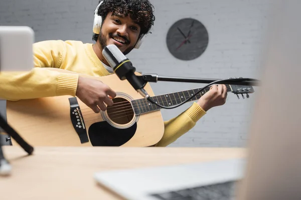 Transmisor indio sonriente en auriculares inalámbricos tocando la guitarra acústica durante la actuación cerca del micrófono y el ordenador portátil borroso en la mesa en el estudio - foto de stock