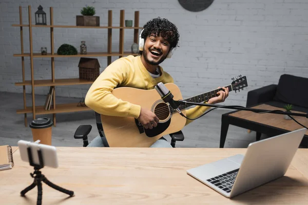 Despreocupado joven podcaster indio en auriculares inalámbricos tocando la guitarra acústica cerca de dispositivos y café para ir en taza de papel y portátil en la mesa en estudio podcast - foto de stock