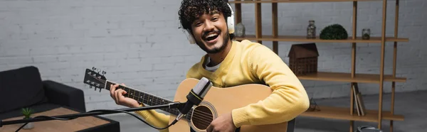 Allegro giovane podcaster indiano in cuffie wireless che suona la chitarra acustica vicino al microfono durante lo streaming in studio podcast, banner — Foto stock
