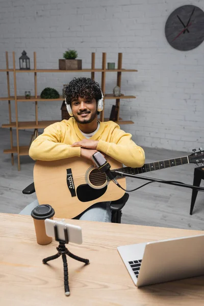 Sonriente podcaster indio en auriculares mirando a la cámara mientras sostiene la guitarra acústica cerca de dispositivos, micrófono y café para ir en estudio podcast - foto de stock