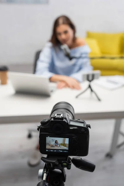 Screen of digital camera on tripod standing near blurred brunette blogger talking at microphone near laptop on table during stream in studio — Stock Photo