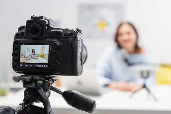 Digital camera on tripod standing near blurred happy female podcaster using devices and microphone on wooden table during stream in podcast studio — Stock Photo