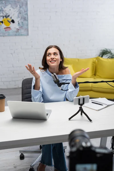 Smiling brunette podcaster looking away while talking near microphone, devices, coffee to go and notebook on wooden table during stream in studio — Stock Photo