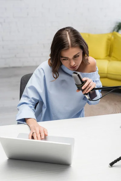Brünette Moderatorin in blauer Bluse, die am Mikrofon spricht und einen verschwommenen Laptop benutzt, während sie beim Online-Stream im Podcast-Studio neben dem Tisch sitzt, mit gelbem Sofa auf verschwommenem Hintergrund — Stockfoto