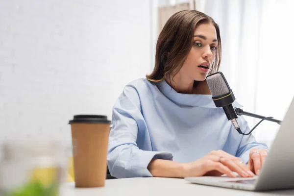 Brunette emissora falando no microfone e usando laptop borrado perto de café para ir em copo de papel na mesa durante o fluxo em estúdio de podcast — Fotografia de Stock