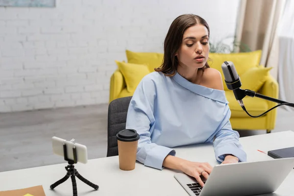 Brunette broadcaster in blouse using laptop near microphone, smartphone on tripod, takeaway coffee and notebook on table during stream  in studio — Stock Photo