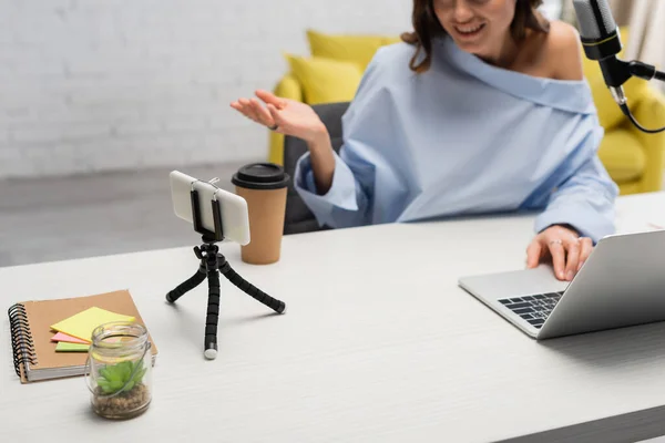 Обрезанный вид размытой брюнетки вещатель говорит рядом со смартфоном и с помощью ноутбука рядом с микрофоном, кофе, чтобы пойти и ноутбук на столе в студии — стоковое фото
