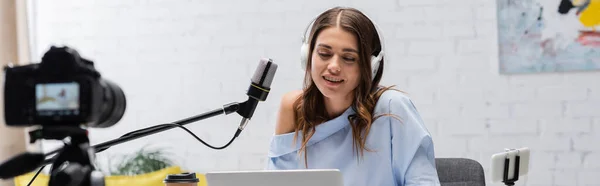 Positive brunette broadcaster in headphones talking during stream near microphone, gadgets and coffee to go next to digital camera in podcast  studio, banner — Stock Photo