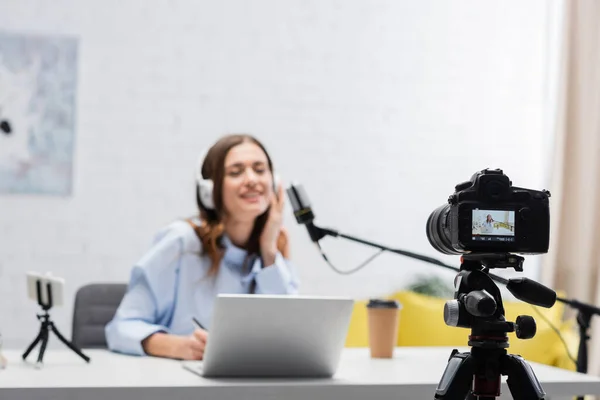 Digital camera near blurred brunette broadcaster in headphones talking during stream near laptop and smartphone on table in podcast studio — Stock Photo
