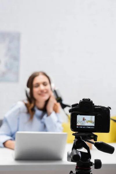 Digital camera on tripod near blurred brunette broadcaster in headphones using laptop near microphone during stream in podcast studio — Stock Photo