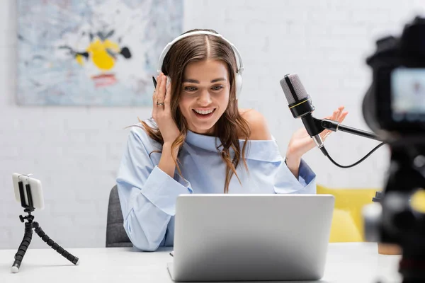 Carefree brunette podcaster in headphones talking and looking at laptop near microphone and blurred digital camera during stream in studio — Stock Photo