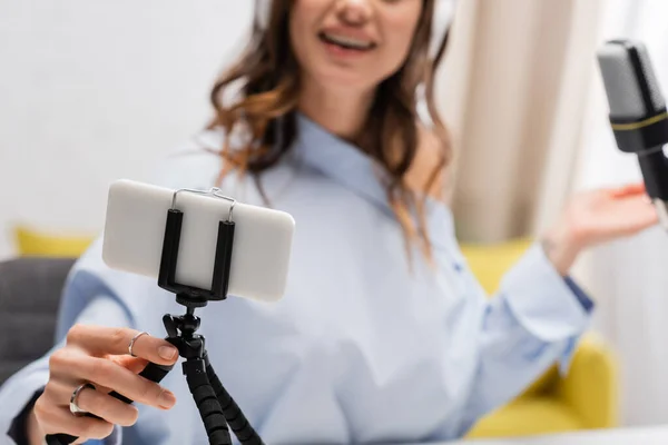 Cropped view of blurred brunette broadcaster in blouse holding smartphone on tripod near microphone during stream in podcast studio — Stock Photo