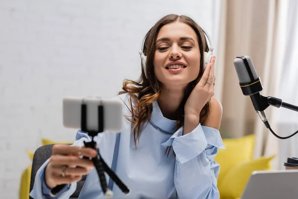 Smiling brunette broadcaster in headphones holding blurred smartphone on tripod near microphone and laptop during stream in podcast studio — Stock Photo