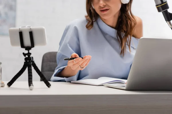 Cropped view of brunette podcaster in blur blouse talking during stream near laptop, smartphone on tripod and microphone during stream in studio — Stock Photo