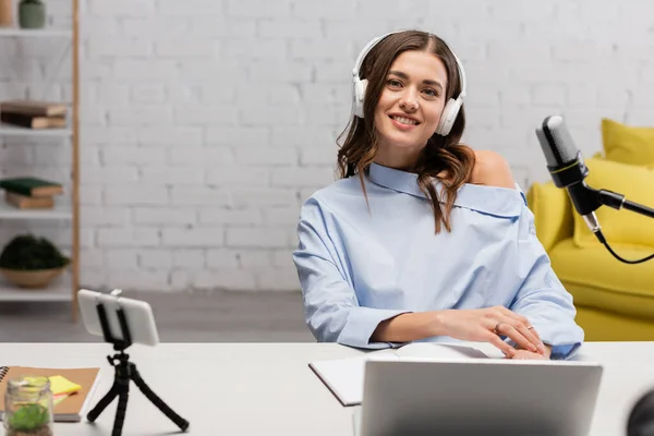 Smiling brunette broadcaster in blue blouse and wireless headphones looking at camera near microphone, gadgets and notebooks in studio — Stock Photo