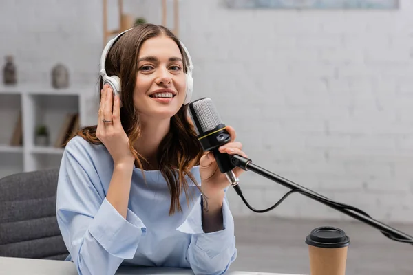Sonriente podcaster morena en auriculares inalámbricos tocando el micrófono y mirando a la cámara cerca del café para ir en taza de papel durante la corriente en el estudio - foto de stock