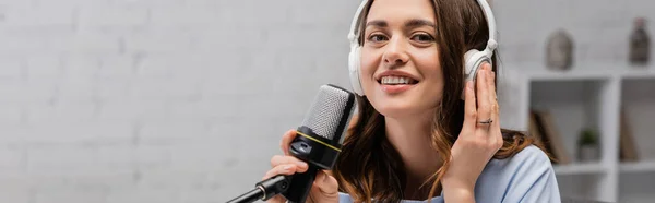 Morena podcaster sonriendo y tocando auriculares inalámbricos sosteniendo micrófono y mirando a la cámara durante la corriente en podcast en el estudio, pancarta - foto de stock