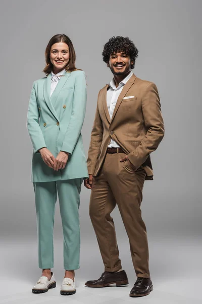 Full length of smiling interracial event hosts in formal wear looking at camera and posing while standing together on grey background — Stock Photo