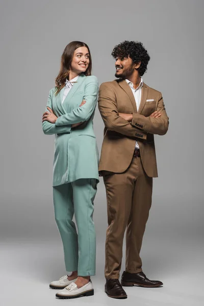Full length of cheerful multiethnic event hosts in suits looking at each other and crossing arms while standing back to back on grey background — Stock Photo