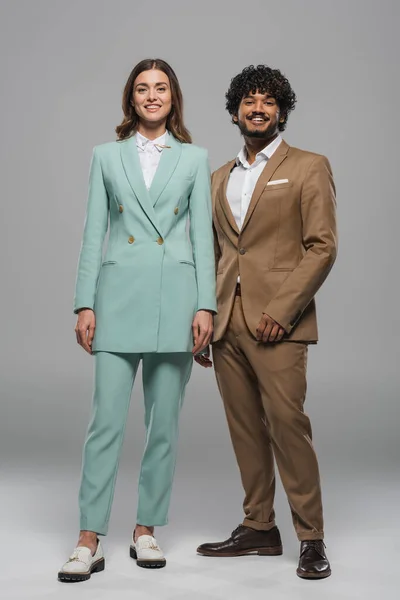 Full length of smiling multiethnic event hosts colleagues in formal wear looking at camera while standing together on grey background — Stock Photo