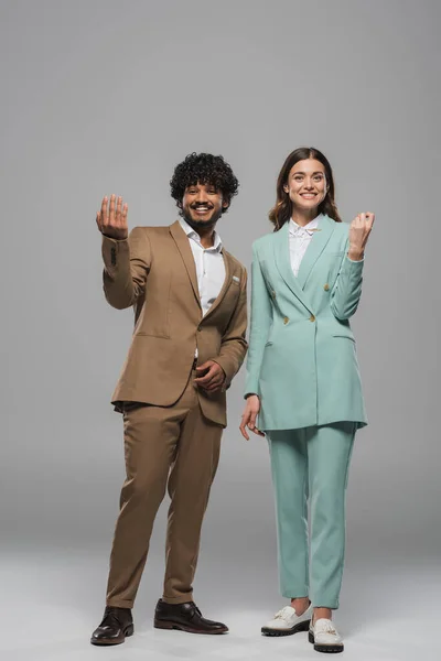Full length of positive and multiethnic event hosts in formal wear gesturing and looking at camera while standing together on grey background — Stock Photo