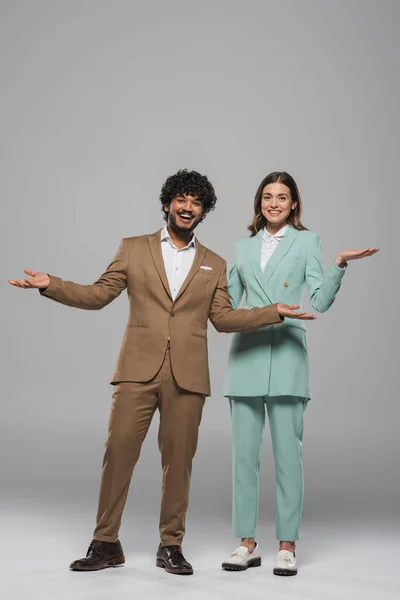 Junge und gut gelaunte indische Moderatoren in formeller Kleidung zeigen mit den Händen, während sie neben Kollegen auf grauem Hintergrund stehen — Stockfoto