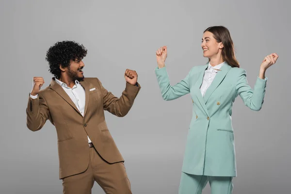 Excited interracial event hosts in formal wear showing yes gesture and looking at each other during holiday while standing isolated on grey — Stock Photo