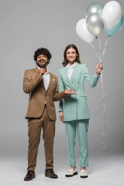 Full length of smiling young indian event host talking at microphone while standing near brunette colleague with colorful balloons on grey background — Stock Photo