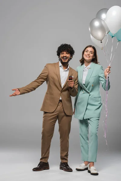 Smiling interracial event hosts in formal wear holding microphone and festive colorful balloons during party while standing together on grey background — Stock Photo