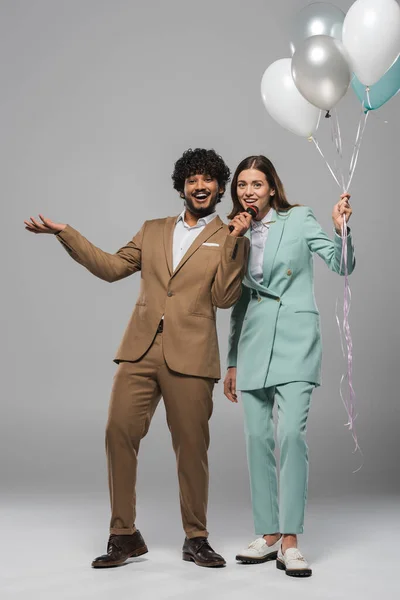 Full length of positive indian event host in formal wear holding microphone near brunette colleague holding festive balloons on grey background — Stock Photo