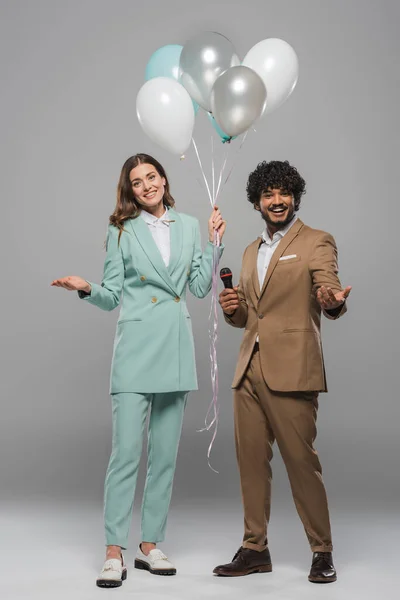 Smiling event host in formal wear holding festive balloons near young indian colleague with microphone while standing together on grey background — Stock Photo