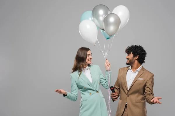 Smiling multiethnic event hosts in formal wear holding microphone and festive balloons while looking at each other and standing isolated on grey — Stock Photo