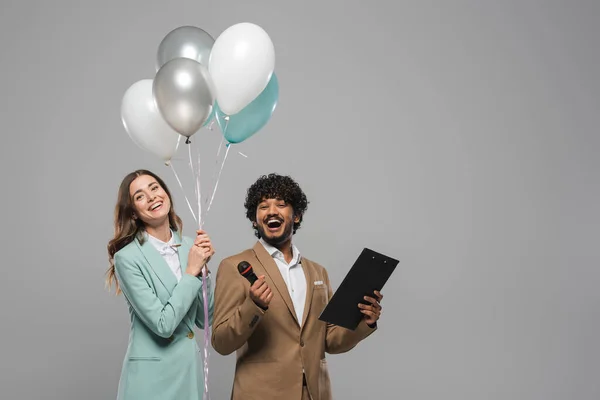 Multiethnic event hosts in formal wear laughing while holding festive balloons and clipboard during performance while standing isolated on grey — Stock Photo