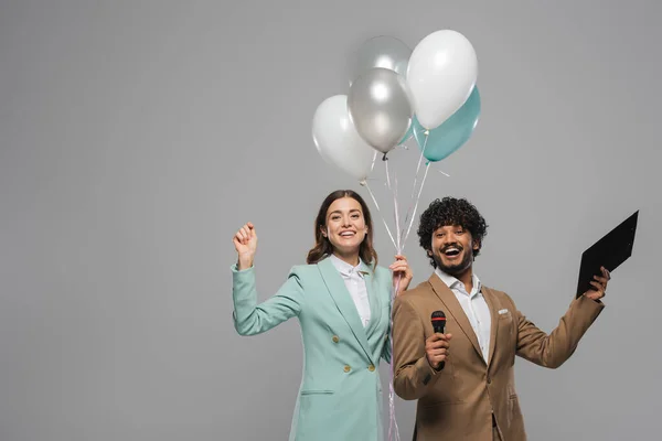 Positive interracial hosts of event in suits holding colorful balloons and microphone during performance while standing isolated on grey with copy space — Stock Photo