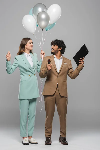 Full length of smiling indian event host in formal wear holding microphone and clipboard near brunette colleague with balloons on grey background — Stock Photo