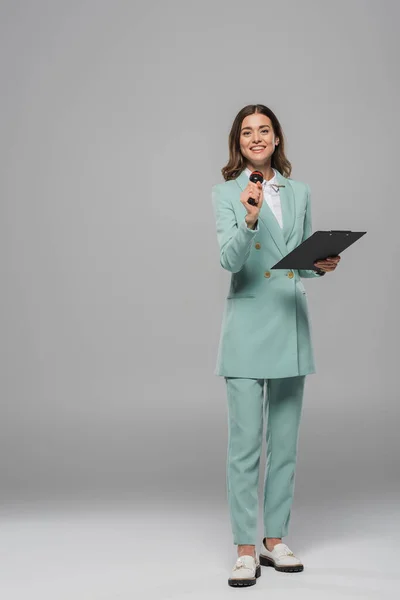 Lächelnder und brünetter Moderator in blauem Anzug, der Mikrofon und Klemmbrett in die Kamera hält und auf grauem Hintergrund steht — Stockfoto