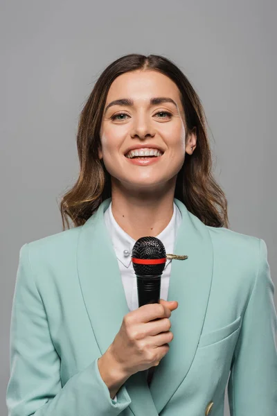 Portrait of smiling brunette event host in blue formal wear talking at wireless microphone and looking at camera while standing isolated on grey — Stock Photo