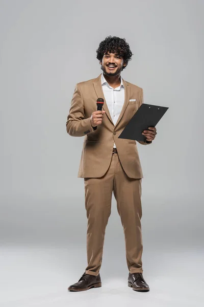 Повна довжина усміхненого ведучого індійського заходу в коричневому формальному одязі, що тримає мікрофон та буфер обміну, стоячи та посміхаючись на сірому фоні — стокове фото