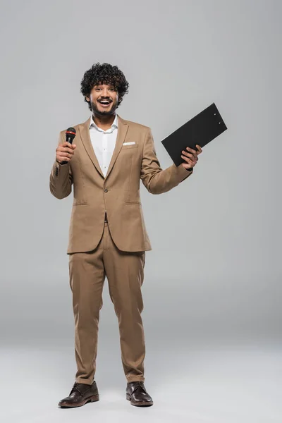 Full length of smiling indian event host in brown suit holding wireless microphone and clipboard during holiday while standing on grey background — Stock Photo