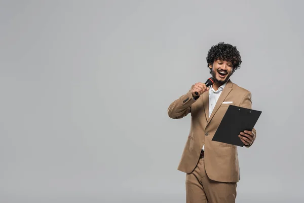 Young and overjoyed indian event host in suit talking at microphone and looking at clipboard during party while standing isolated on grey with copy space — Stock Photo