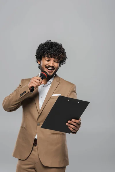 Young indian host of event in formal wear smiling while talking at microphone and holding clipboard during party and standing isolated on grey — Stock Photo