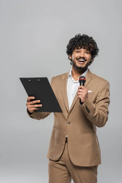 Cheerful and curly young indian host of event holding microphone and clipboard while looking at camera during celebration isolated on grey — Stock Photo