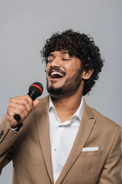 Porträt eines fröhlichen indischen Moderators mit geschlossenen Augen in formeller Kleidung, der während des Auftritts am Mikrofon spricht, während er isoliert auf grau steht — Stockfoto